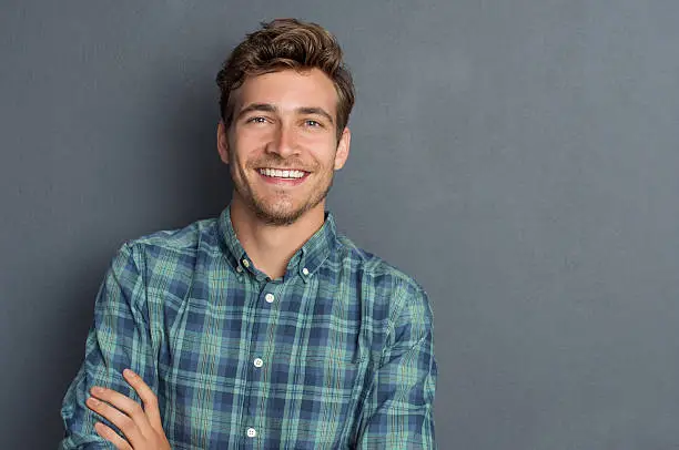 Young handsome man leaning against grey wall with arms crossed. Cheerful man laughing and looking at camera with a big grin. Portrait of a happy young man standing with crossed arms over grey background.