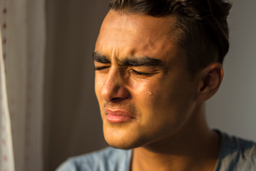 Portrait of crying young man closing his eyes near window. Image taken at home near window. Worried young man crying in pain. He is closing his eyes, one teardrop is falling into the down slowly... Horizontal composition.