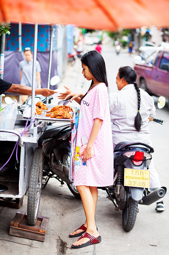 Bangkok, Thailand - June 30, 2016: A thai girl wearing chemise and nightdress is buying food at street food market stall near Chandrakasem university. In background a thai woman is on motorcycle.