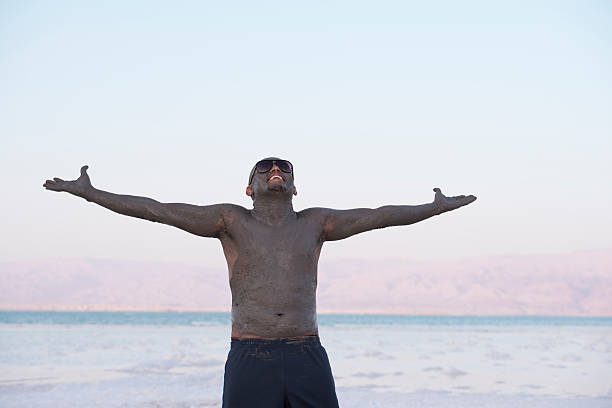 Thanks to Dead Sea. Waist up portrait of handsome man with wide open raised hands. Man wearing sunglasses, standing on Dead Sea beach. Man smeared with healing mud. Body care treatment by natural mineral mud sourced from the salty lake. people covered in mud stock pictures, royalty-free photos & images