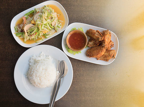Cucumber salad (Som Tam),Fried chicken wings,cooked rice,food thai style.Cooked rice in bowl with spoon and dishcloth on old wooden table