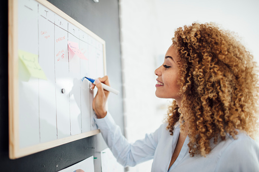 Side view of latin businesswoman standing near whiteboard and writing. She is making schedule for next business week.