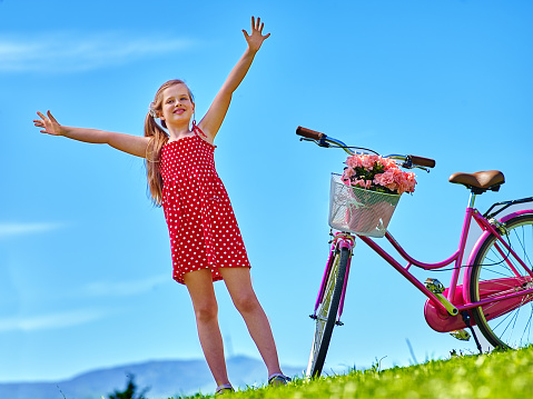 Bikes cycling girl. Child girl wearing red polka dots dress rides bicycle with flowers basket. Mountains and blu sky on background.