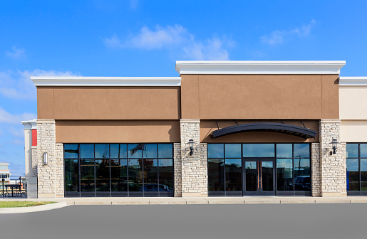 The front view of a new commercial retail space in a strip mall ready to rent. The photograph includes showing the sidewalk and driveway.  Blue sky and clouds are in the background. The building is located in East Peoria, IL