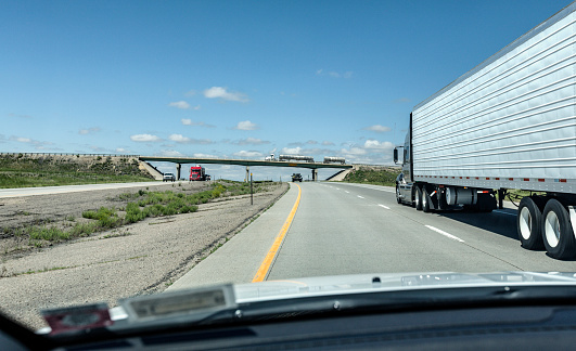 Looking over the dashboard through the windshield of a speeding car while passing a long eighteen wheeler freight hauling trailer truck on a sunny summer uphill section of the Interstate 80 expressway highway near Green River, Wyoming, USA. Several other trucks are visible driving on the road in both directions, and a tandem tanker truck is hauling liquid freight across the highway overpass bridge at the top of the hill.