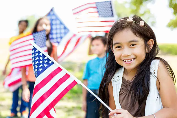 Adorable elementary age Hispanic little girl smiles and giggles as she waves an American flag at Independence Day parade or party. Her friends are behind her also waving flags.