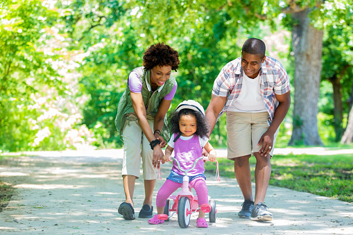 Happy African American mother and father teaching their toddler daughter to ride a tricycle. She is smiling and is wearing a pink helmet. The tricycle is also pink. There are many trees behind them on the path.