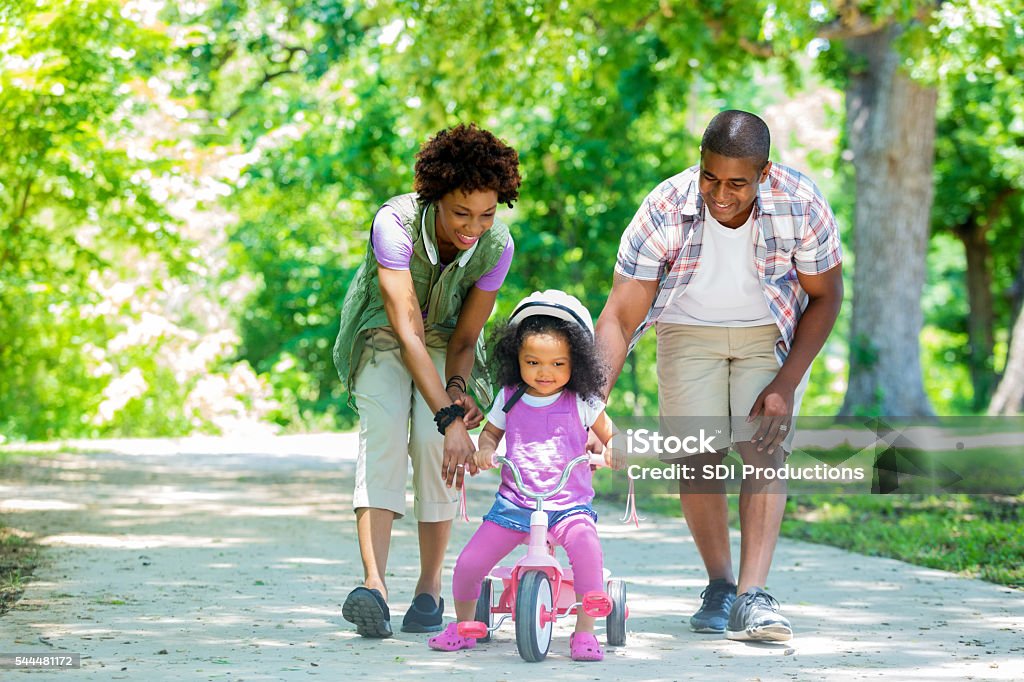 Happy Mother and Father teaching toddler daughter to ride tricycle Happy African American mother and father teaching their toddler daughter to ride a tricycle. She is smiling and is wearing a pink helmet. The tricycle is also pink. There are many trees behind them on the path. Child Stock Photo