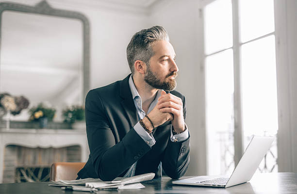 Businessman working from home Businessman working from home sitting with a pile of loose paperwork and a laptop computer staring out of the window with a pensive expression business man looking away stock pictures, royalty-free photos & images