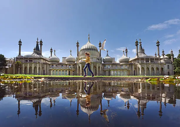 A teenage girl reflected from across a pond - runs pursued by a seagull past the seaside town's landmark Georgian Royal Pavilion with it's famous domes and minarets