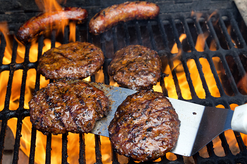 Juicy hamburgers ready to be turned by a metal spatula over an outdoor charcoal grate  with a background of perfectly seared brats being licked by the flames.