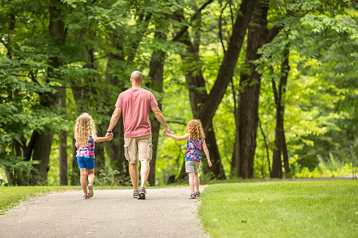 Rear view of two young girls walking along a park trail holding Dad's hands. The older sister is on the left with curly blonde hair, Dad is in the middle, and the younger sister is on the right with curly red hair. Taken on a summer day in Minnesota.