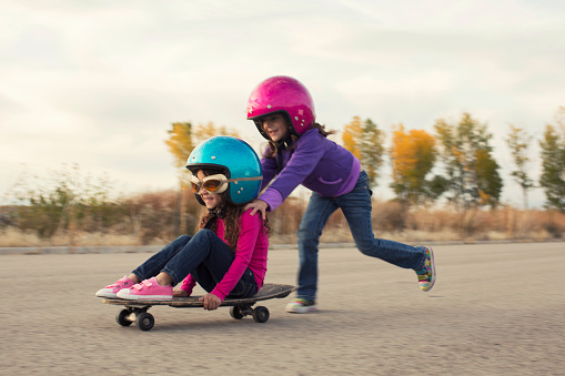 Two young girls are racing on skateboard on a rural road in Utah. They are wearing helmets, and are smiling while they are going fast. One girl is pushing the other and they make a great team.