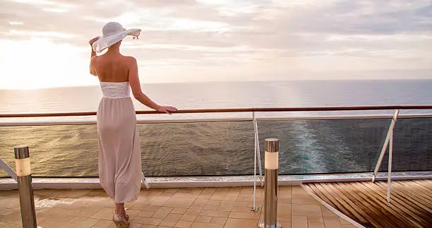 Rear view of young woman looking at sea. Female is standing at railing in cruise ship against sky. She is wearing white dress and sun hat.