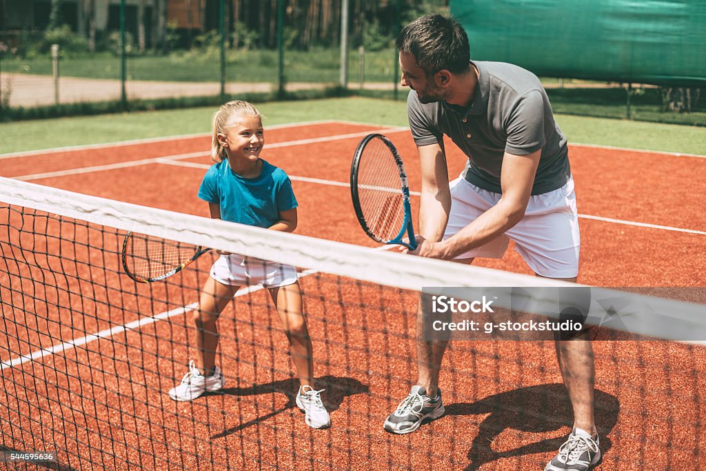 Tennis training. Cheerful father in sports clothing teaching his daughter to play tennis while both standing on tennis court Tennis Stock Photo