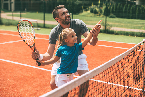 Cheerful father in sports clothing teaching his daughter to play tennis while both standing on tennis court