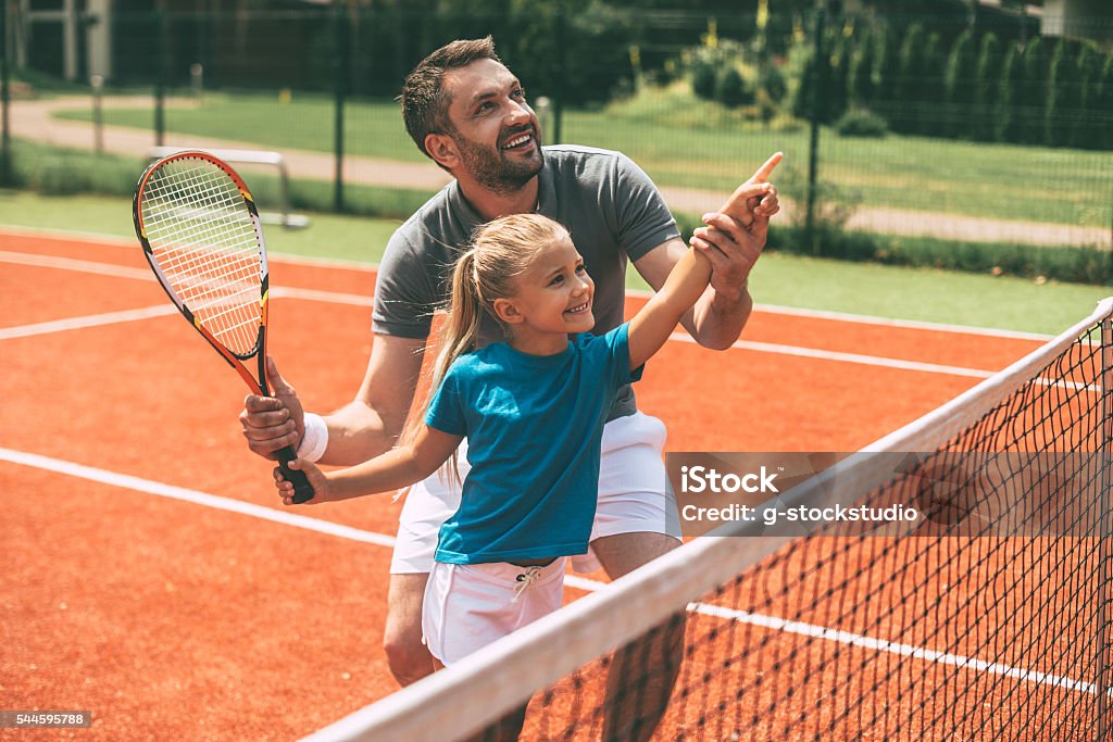 Tennis is fun when father is near. Cheerful father in sports clothing teaching his daughter to play tennis while both standing on tennis court Tennis Stock Photo