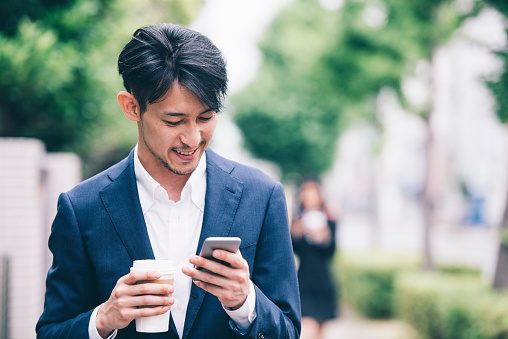 Businessman using mobile phone while walking outdoors. He is holding cup of coffee in other hand
