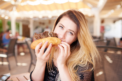lifestyle shot of young woman eating steak sandwich and biting from meat, looking at camera and being hungry.