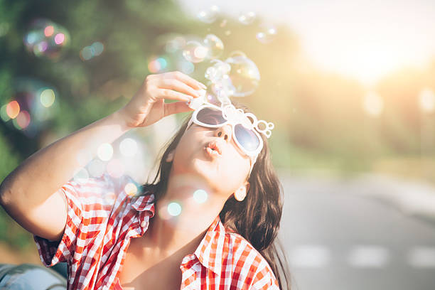 Young woman with sunglasses ans checkered shirt blowing soap bubbles on street. Focus on foreground. Looking up.