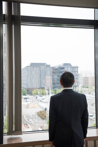 Mature man in suit jacket by window, rear view of man in office looking out towards buildings