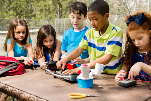 Group of multi-ethnic, elementary age children collaborate on a 'robot' they created using tools, objects they found at home. Imagination, creativity, inspiration for these little engineers!  Park or schoolyard setting.