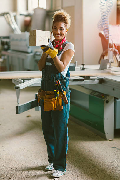 Latina Carpenter Standing In Her Workshop Latina carpenter standing in her workshop. She is carry wooden plank and smiling. Wearing protective gloves. woman wearing tool belt stock pictures, royalty-free photos & images