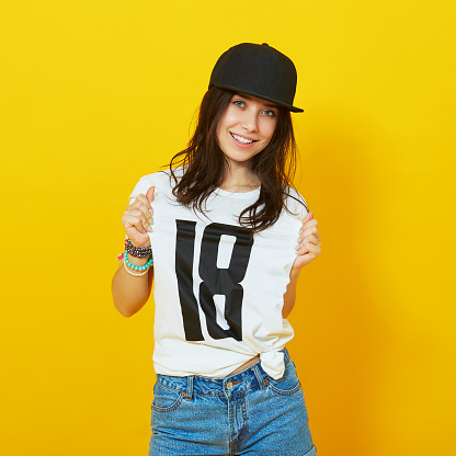 Young beautiful woman posing with white t-shirts with 18 text on it. Girl turned 18 concept. Eighteen years old teenage girl pointing to a tshirt over a bright yellow background.