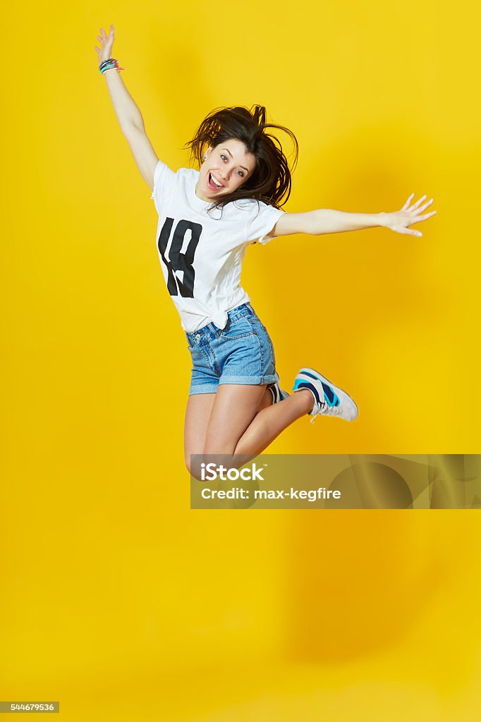 Girl showing her tongue and peace gesture Cheerful hipster girl jumpig with hands spread wide over yellow background. Young woman in jump. Jumping Stock Photo