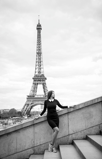 Young woman in a black dress is standing gracefully on old stairs near the Eiffel Tower in Paris and enjoying the atmosphere of the beautiful city. She seems greatly relaxed and elegant. Great composition with the Eiffel Tower in the background. Copy space available. Black and white image.