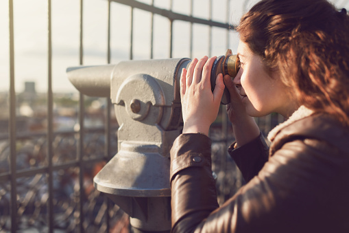 Young caucasian woman looking at the city through a coin operated binoculars, close-up.
