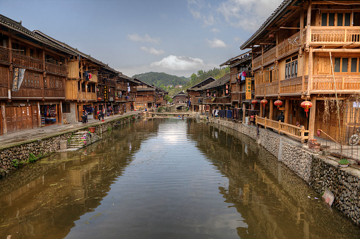 Zhaoxing Dong Village, Guizhou Province, China - April 8, 2010: Rural River with cobblestone seafront promenade and wooden houses with trade rows on both shores,  village ethnic minorities, sunny day