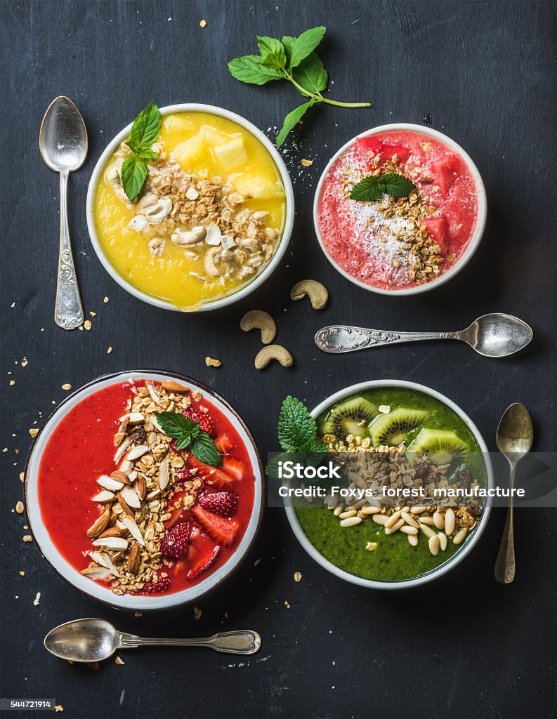 Healthy summer breakfast concept. Colorful fruit smoothie bowls with nuts Healthy summer breakfast concept. Bowls with colorful fruit smoothie with nuts, oat granola and mint leaves served with silver spoons on black background . Red, yellow, green, pink. Top view, vertical composition Almond Stock Photo