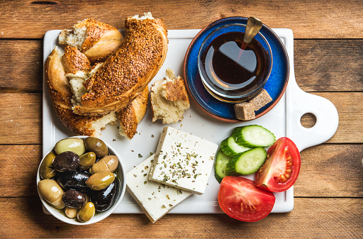 Turkish traditional breakfast with feta cheese, vegetables, olives, simit bagel and black tea on white ceramic board over wooden background. Top view, horizontal composition