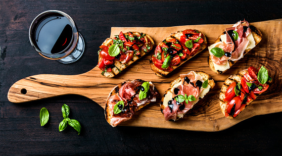 Brushetta set for wine. Variety of small sandwiches with prosciutto, tomatoes, parmesan cheese, fresh basil and balsamic creme served with glass of red wine on rustic wooden board over dark background, top view