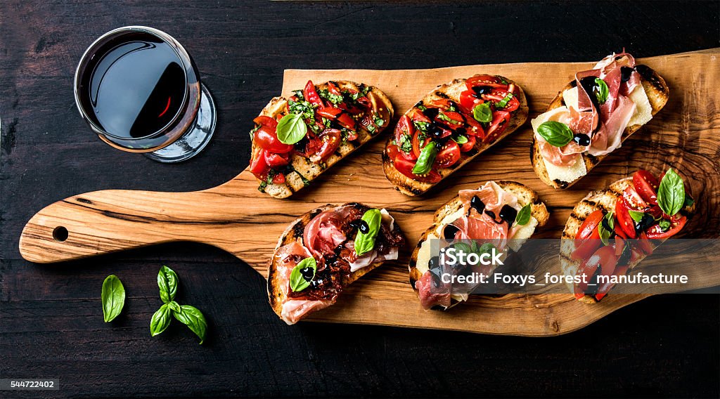 Brushetta set and glass of red wine. Small sandwiches with Brushetta set for wine. Variety of small sandwiches with prosciutto, tomatoes, parmesan cheese, fresh basil and balsamic creme served with glass of red wine on rustic wooden board over dark background, top view Tapas Stock Photo
