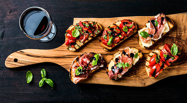 Brushetta set and glass of red wine. Small sandwiches with Brushetta set for wine. Variety of small sandwiches with prosciutto, tomatoes, parmesan cheese, fresh basil and balsamic creme served with glass of red wine on rustic wooden board over dark background, top view appetizer stock pictures, royalty-free photos & images