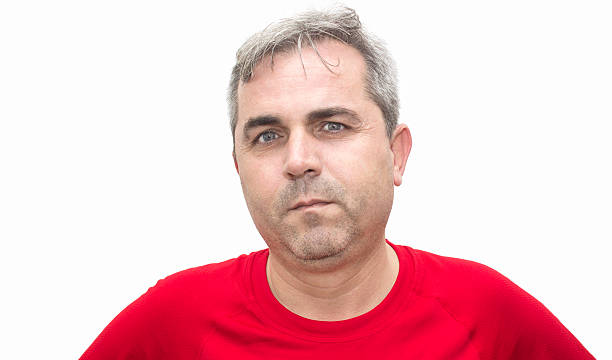 Portrait of mature man looking at camera with blank facial expression over isolated on white. Horizontal compositipon. Studio shot. Relaxed mature man standing in front of white background in causual clothes (he is wearing a red t-shirt) looking at camera. He has got short, white-gray hair and blue eyes. His ethnicity belongs to Turkish ethnicity.