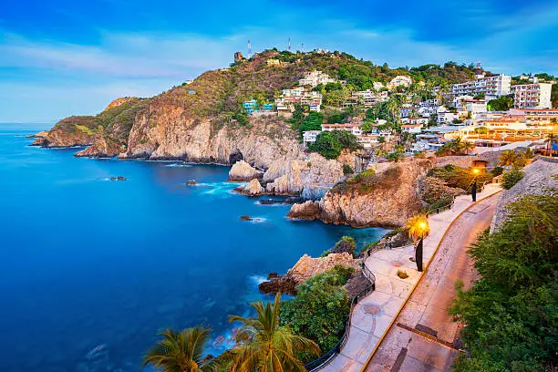 Photo of a rocky coastline with illuminated promenade and the Pacific Ocean in Acapulco, Mexico at dawn.