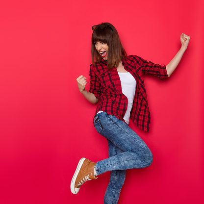 Young woman in red lumberjack shirt, jeans and brown sneakers standing on one leg, flexing muscles, looking over the shoulder and shouting. Three quarter length studio shot on yellow background.