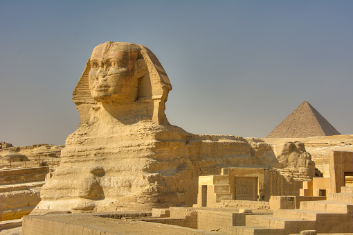 The Great Sphinx of Giza. Monumental limestone statue of a reclining sphinx with a lion's body and a human head (believed to represent the face of the Pharaoh Khephren). One of the pyramids in the background. Giza, Egypt