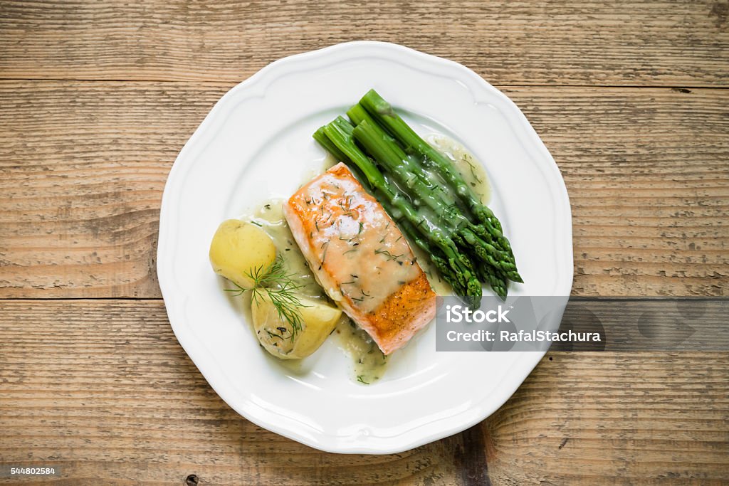 Grilled salmon with boiled potatoes and asparagus. Top view Top view of grilled salmon fillet with potatoes and asparagus garnished with dill sauce arranged on white plate and old wooden table Salmon - Seafood Stock Photo