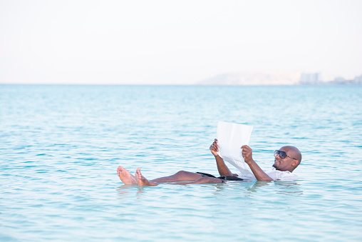 Handsome man reading newspaper while floating in Dead Sea, Israel, Ha Negev.
