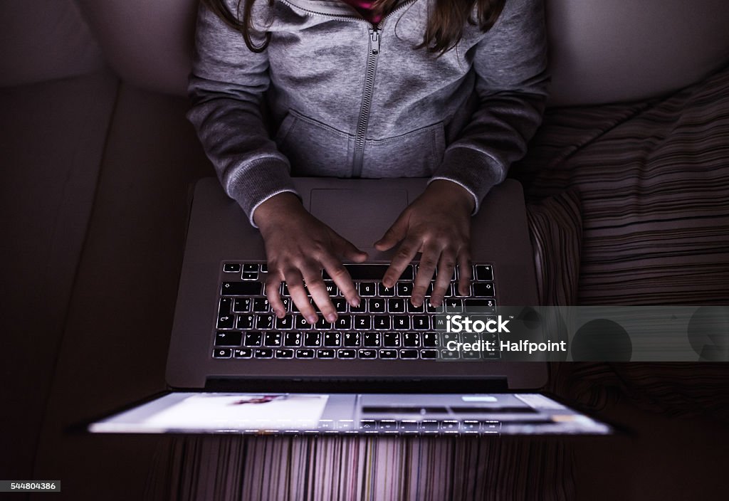 Unrecognizable girl, sitting in a dark, playing with laptop Unrecognizable girl, sitting in a dark, playing with laptop. Child at home, sitting on sofa. Dark Stock Photo