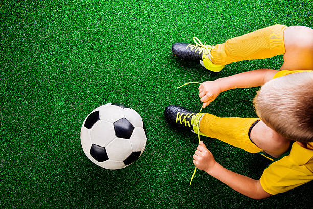 Unrecognizable little football player against green grass, studi Unrecognizable little football player with soccer ball tying shoelaces, against artificial grass. Studio shot on green grass. cleat stock pictures, royalty-free photos & images