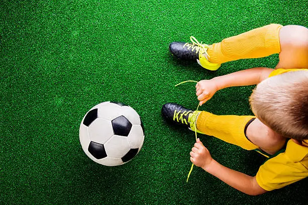 Unrecognizable little football player with soccer ball tying shoelaces, against artificial grass. Studio shot on green grass.