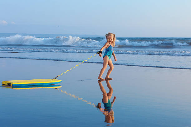 Young surfer girl with bodyboard walks along beach sea surf Little baby girl - young surfer with bodyboard has fun on sea beach. Family lifestyle, people outdoor water sport lessons and swimming activity on summer vacation with child in adventure surf camp. body board stock pictures, royalty-free photos & images
