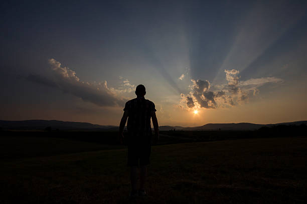 Man standing on the field in front of setting Sun, with crepuscular rays in the sky. Photo is taken with dslr camera and wide angle lens, at the end of summer day in European countryside.