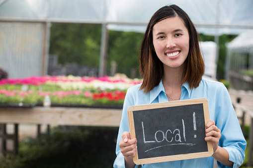 Beautiful Asian young woman holds chalkboard sign with 'Local!' printed on it in local farmers' market or garden center. She is wearing a light blue button down shirt and has dark brown hair.