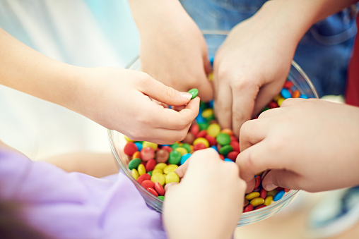 Hands of kids taking multi colored chocolate candies from bowl. Children love sweet food. They eating colorful button-shaped chocolates. Its photo illustrating happy childhood. It is perfect for using it in commercial and advertising photography, reports, books, presentation